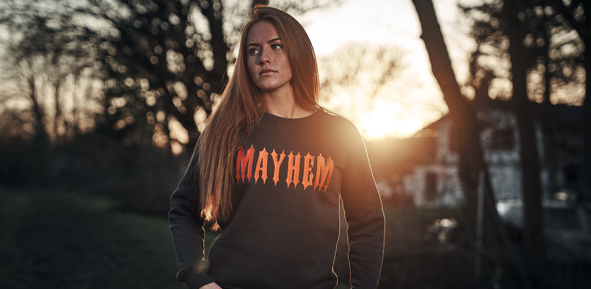 frau mit roten haaren vor sonnenuntergang trägt men of mayhem women crewneck in grau mit orange farbenem stick auf der brust