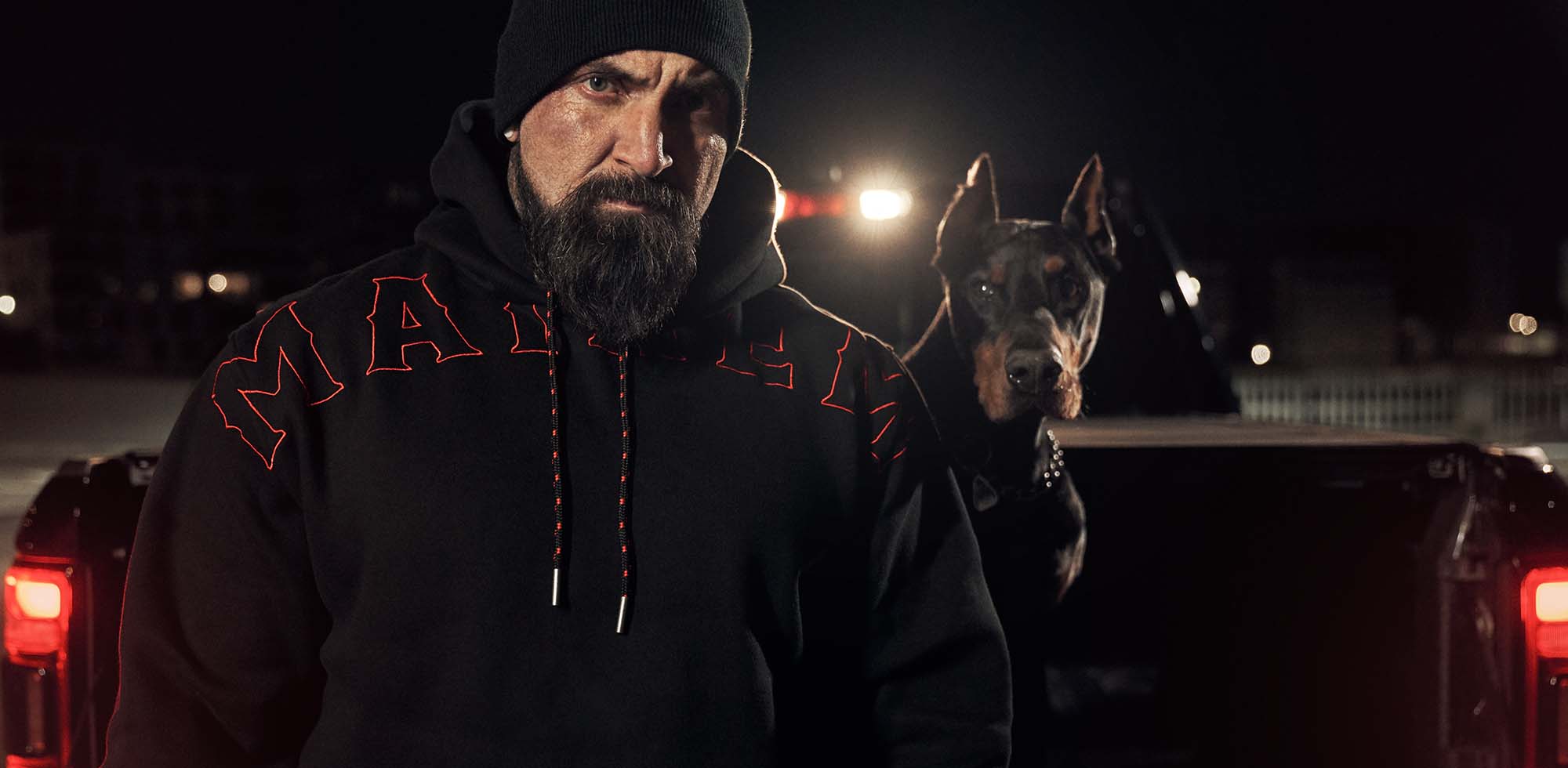 mann bei nacht steht vor pickup truck mit dobermann und trägt men of mayhem crook hoody in schwarz rot