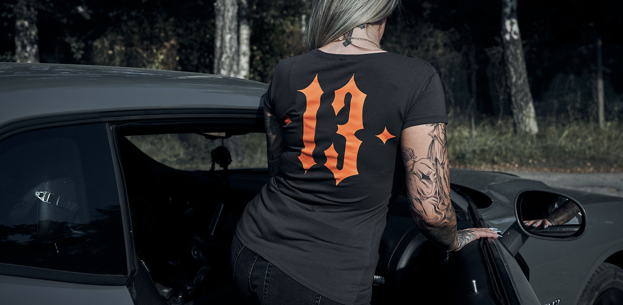 blonde frau mit tattoos steigt ins auto ein und trägt dabei men of mayhem t-shirt classic in grau - zu sehen auf dem rücken ein großer 13 print