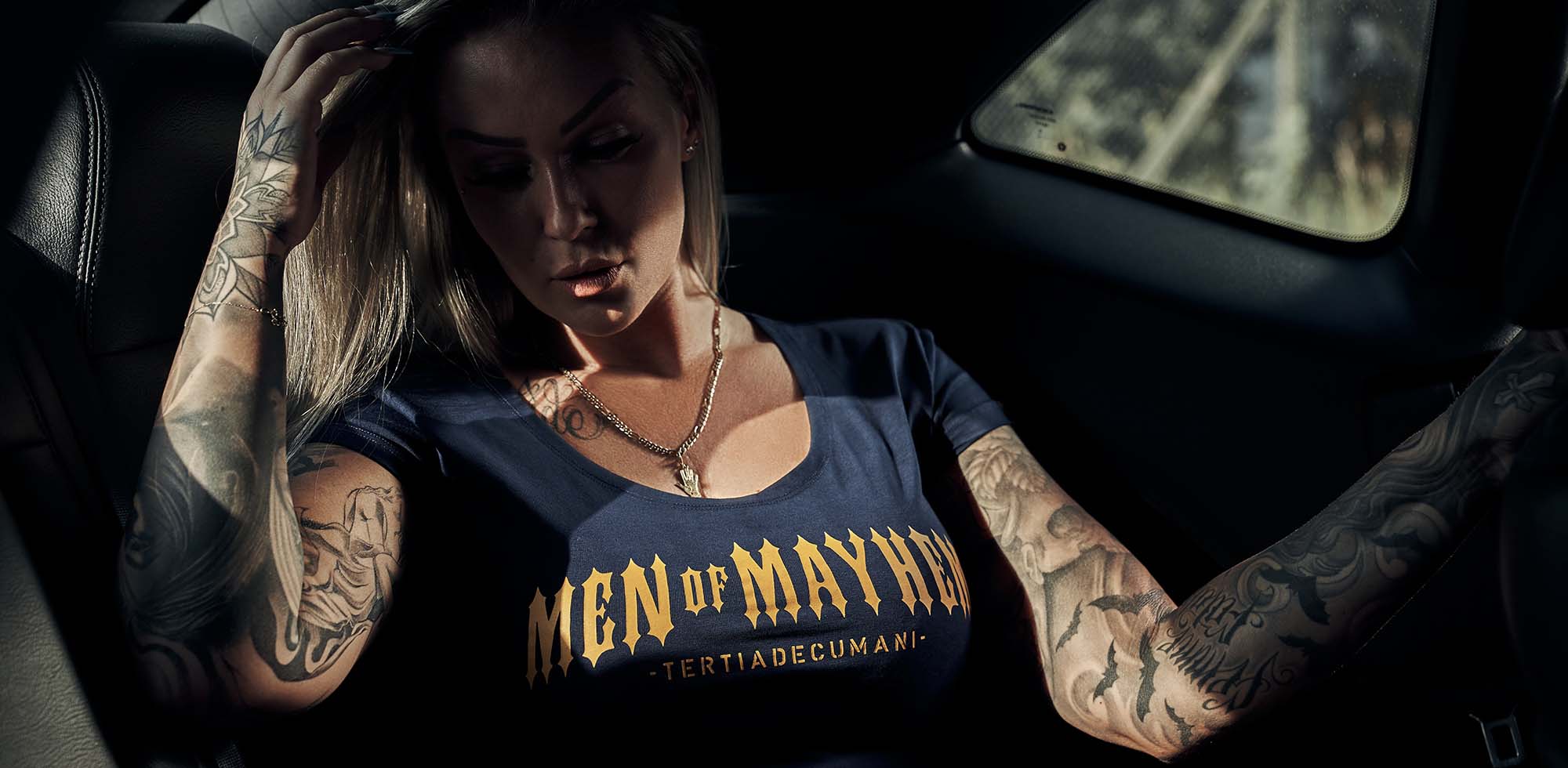 blonde frau mit tattoos sitzt im auto und trägt men of mayhem women classic t-shirt in navy mit gelb/gold print auf der brust