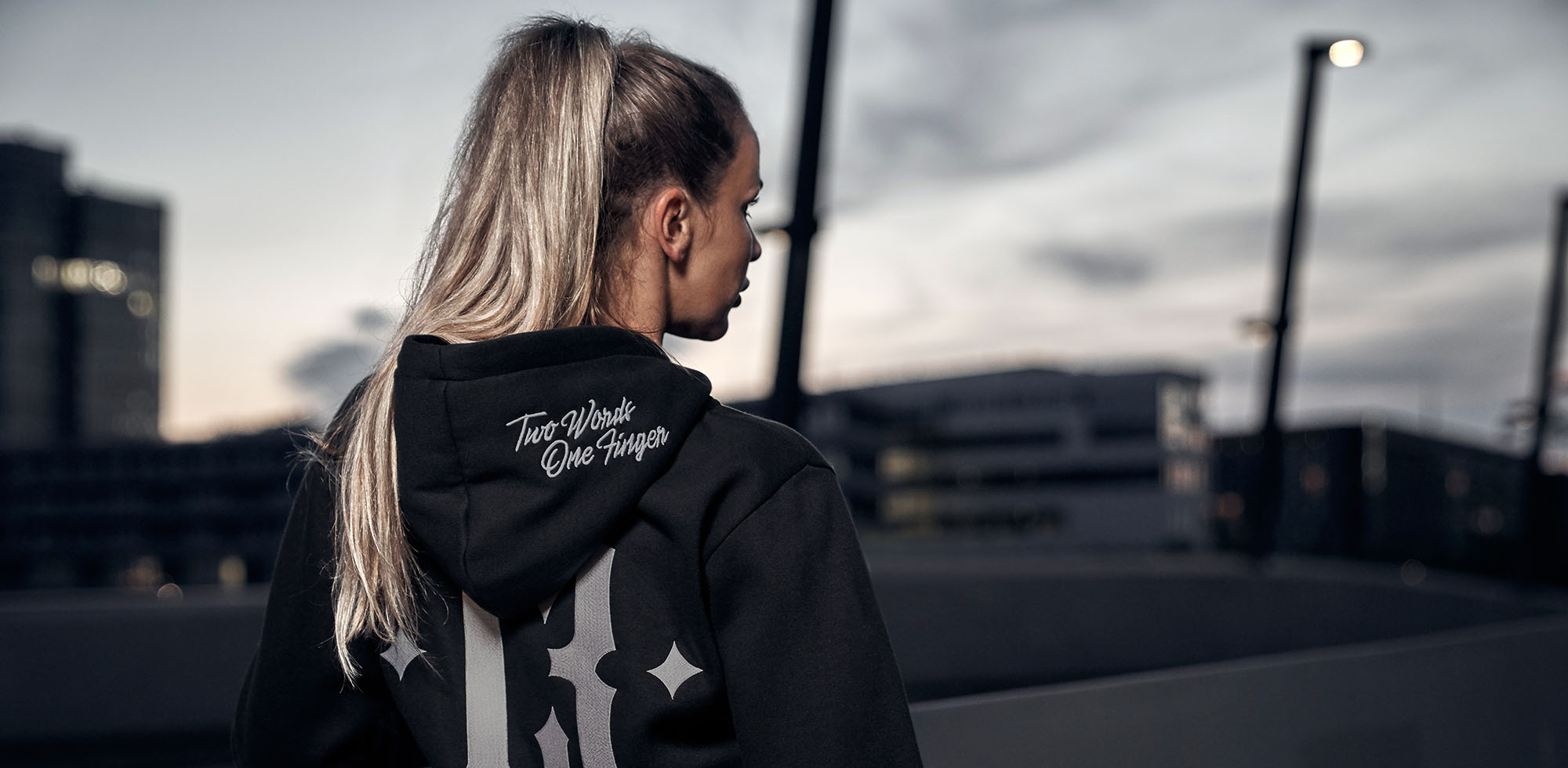 blonde frau steht am hafen bei dämmerung und trägt men of mayhem women zipper T.w.o.f. in schwarz grau mit großer 13 auf dem rücken