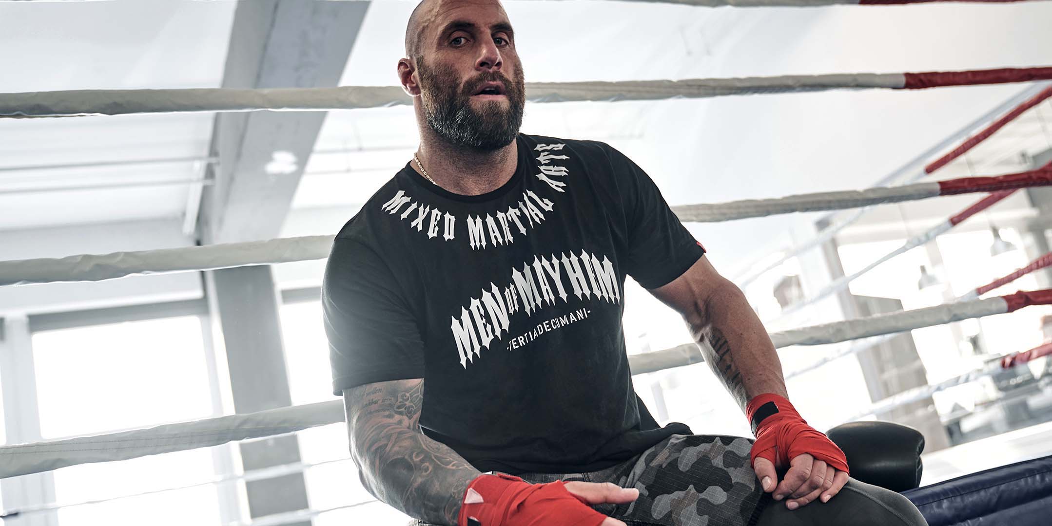 mann sitzend im boxring mit bandagierten händen trägt men of mayhem fight team t-shirt in schwarz weiss