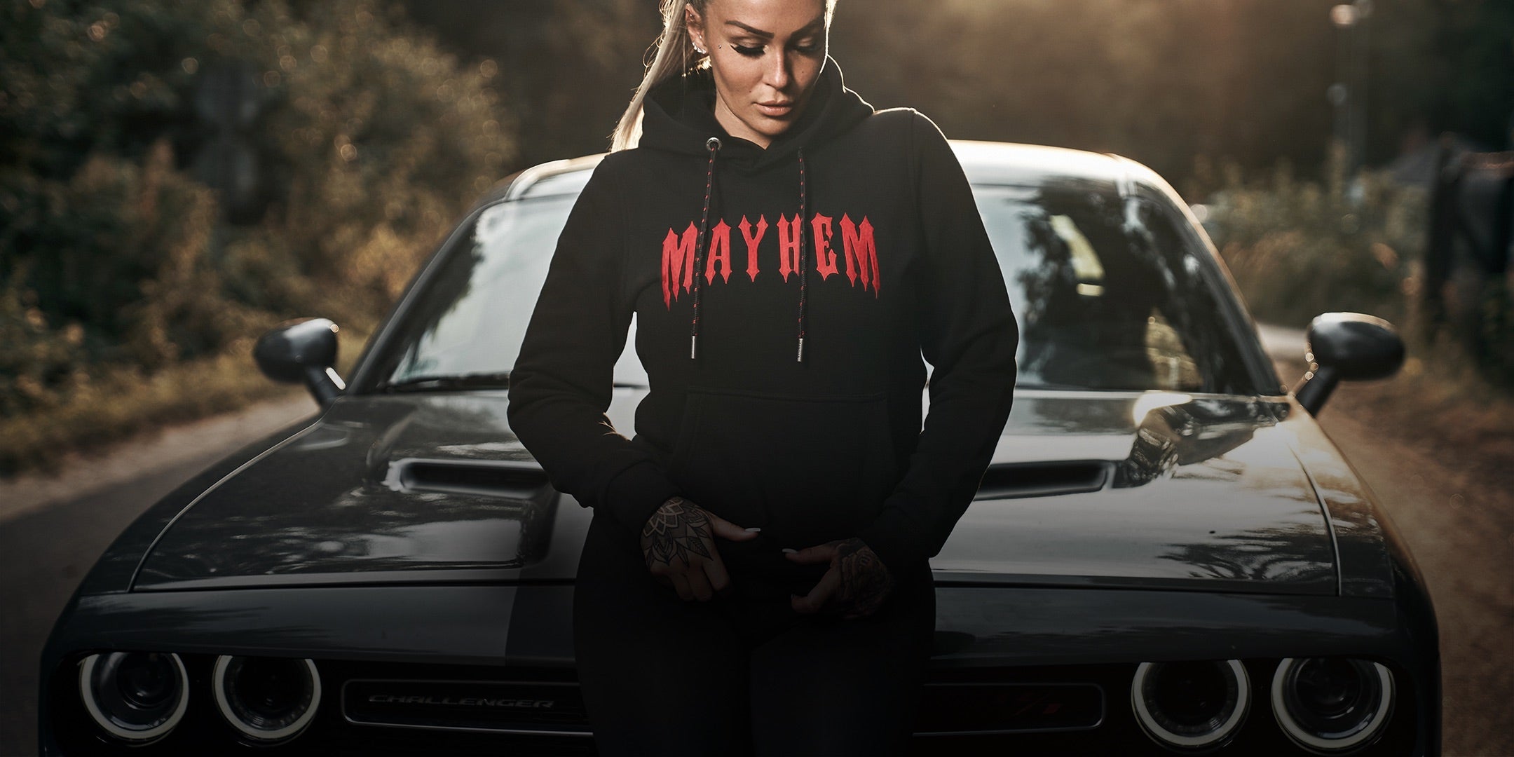 Frau mit Tattoos trägt schwarzen Mayhem Hoodie neben Auto – Women Collection von Men of Mayhem