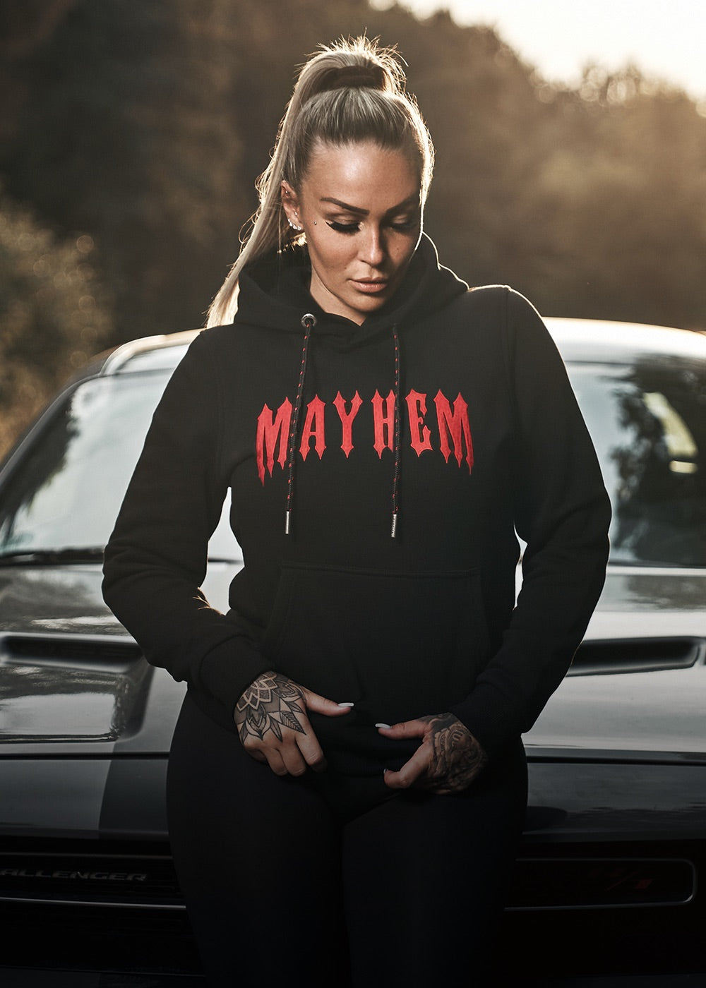 Frau mit Tattoos trägt schwarzen Mayhem Hoodie neben Auto – Women Collection von Men of Mayhem