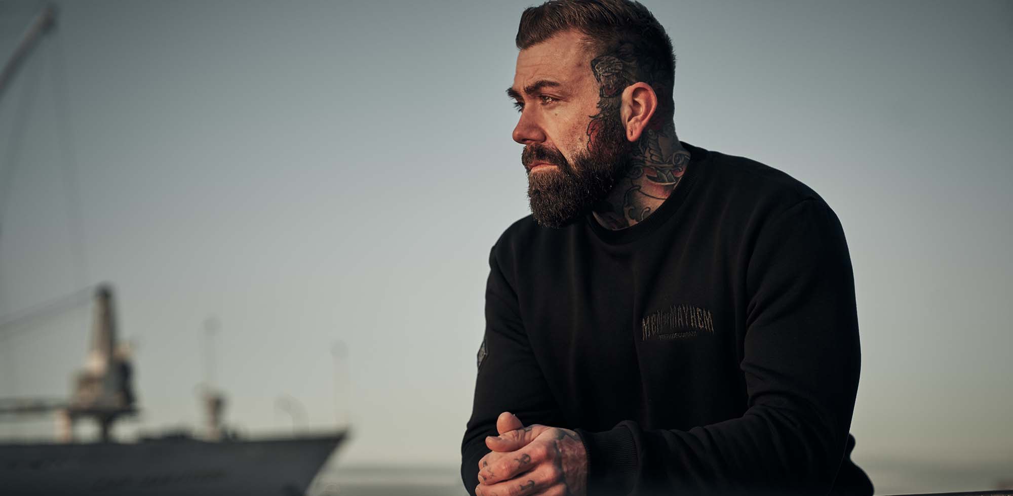 mann stehend am hafen trägt men of mayhem crewneck pullover in schwarz mit men of mayhem stick auf der brust