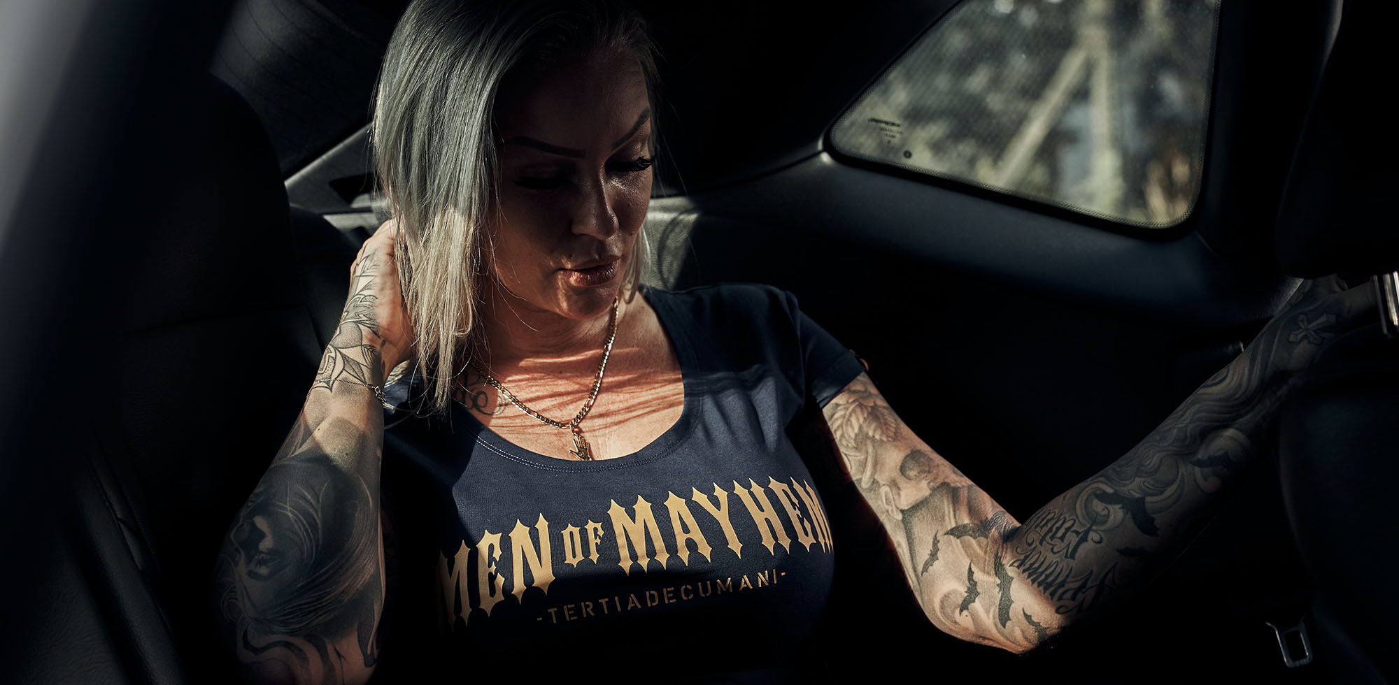 blonde frau mit tattoos sitzt im auto und trägt men of mayhem women classic t-shirt in navy mit gelb/gold print auf der brust