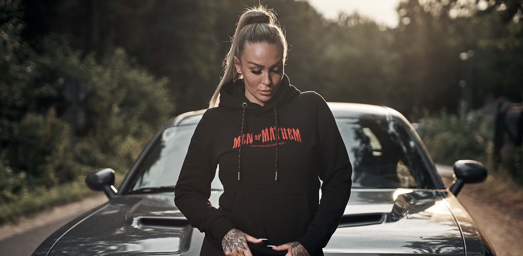 frau mit blonden haaren steht vor muscle car und trägt men of mayhem hoody in schwarz rot